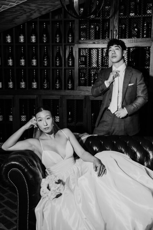 Bride in a white dress sits on a couch, while groom adjusts his tie, with wine bottles behind them—capturing the intimate charm of a Barrel House wedding.