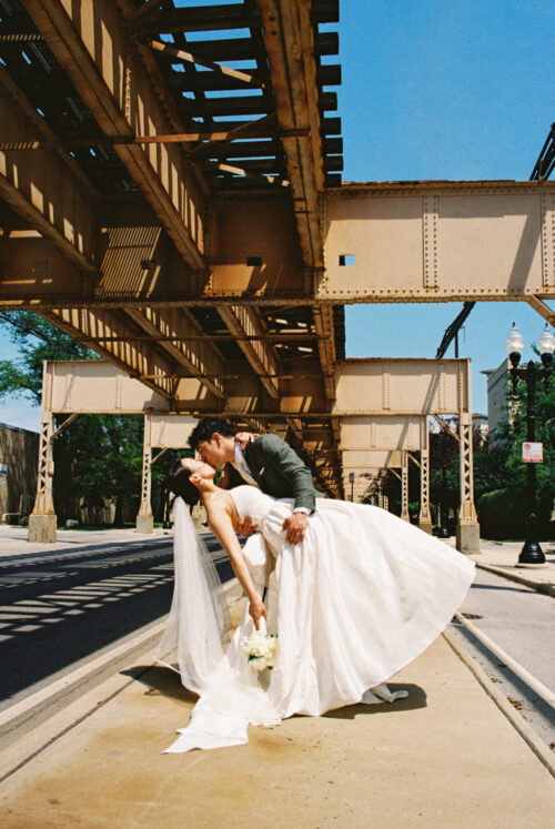 A groom dips and kisses his bride under a metal train bridge, both smiling in wedding attire during their unforgettable Barrel House wedding.