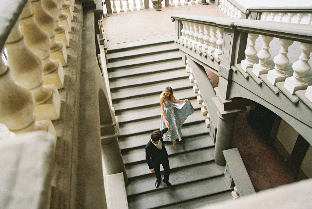 A woman in a blue dress and a man in a suit walk down a grand staircase with stone railings in Lucca, Italy