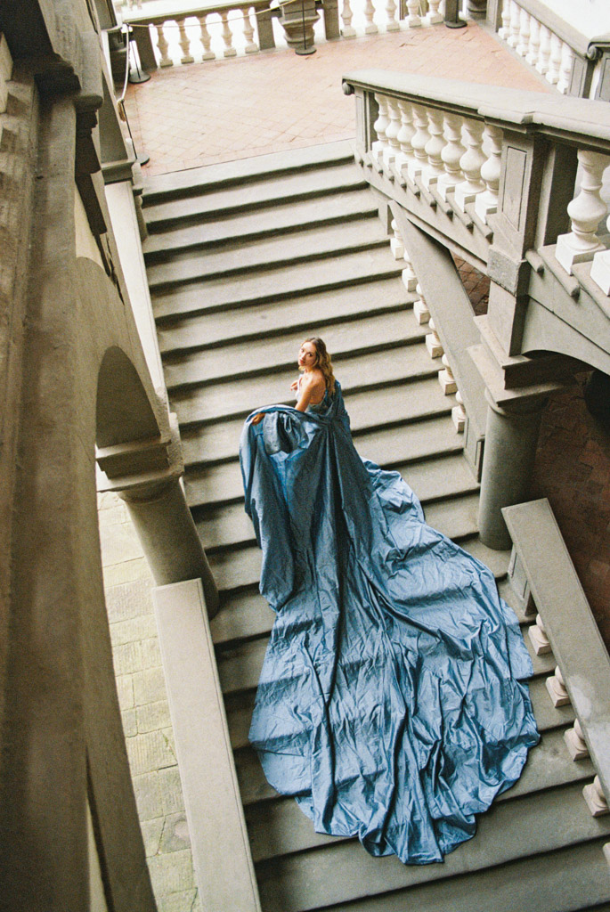 Woman in a flowing blue gown stands on grand stone stairs, looking back towards the camera, during the elegance of a Lucca, Italy engagement session