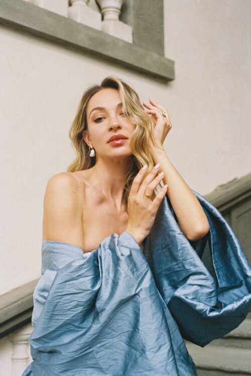 Woman with wavy blonde hair poses in a blue off-shoulder dress on a staircase in Lucca, Italy