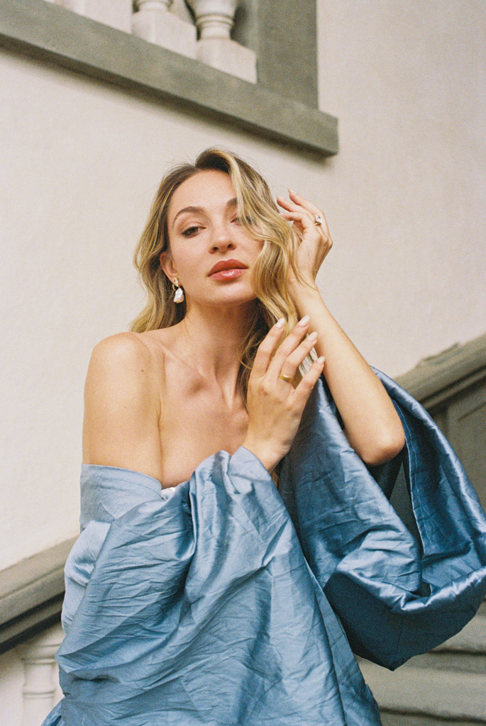 Woman with wavy blonde hair poses in a blue off-shoulder dress on a staircase in Lucca, Italy