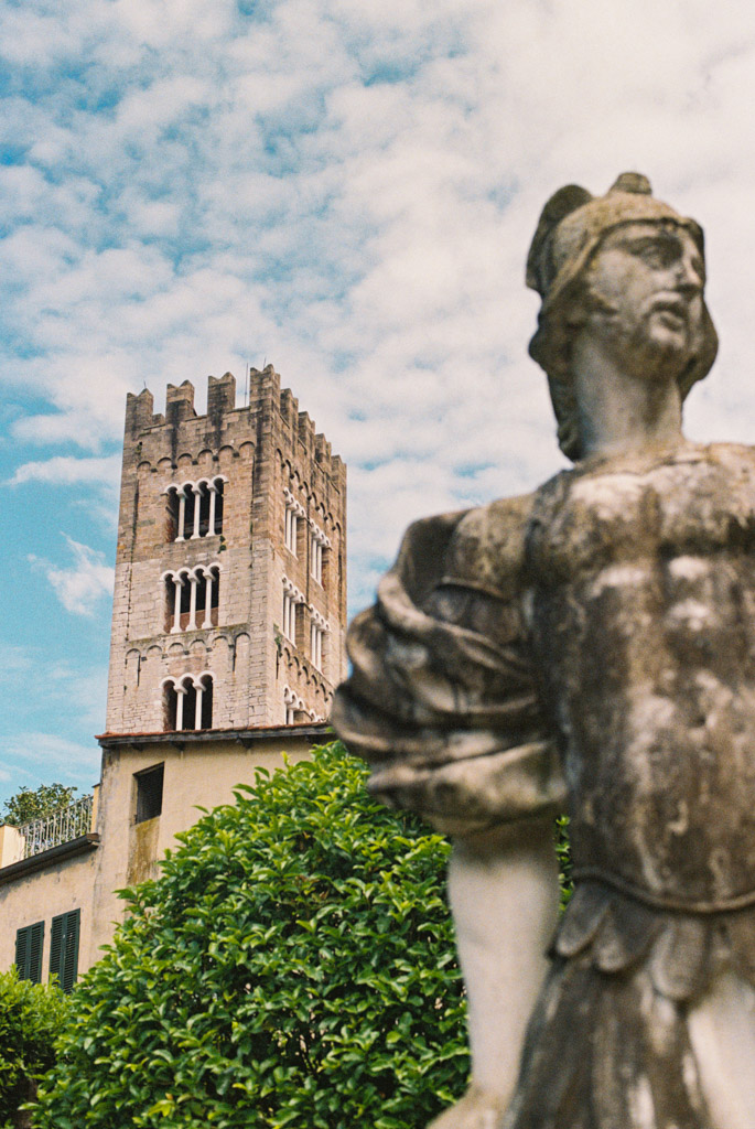 Film photo of stone statue in Lucca, Italy with tall brick bell tower and green trees under a partly cloudy sky in background