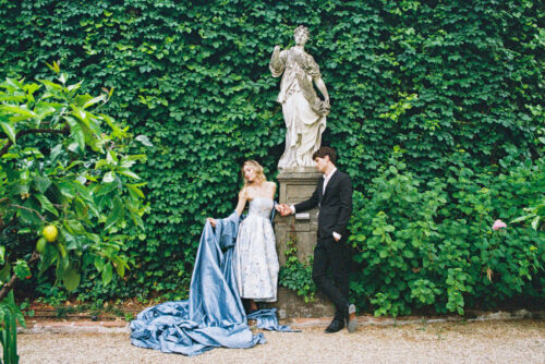 A woman in a blue gown and a man in a suit stand by a statue, surrounded by lush green foliage, during their Lucca engagement session