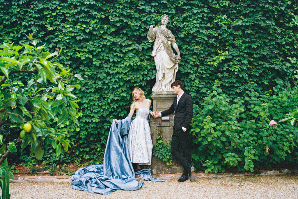 A woman in a blue gown and a man in a suit stand by a statue, surrounded by lush green foliage, during their Lucca engagement session