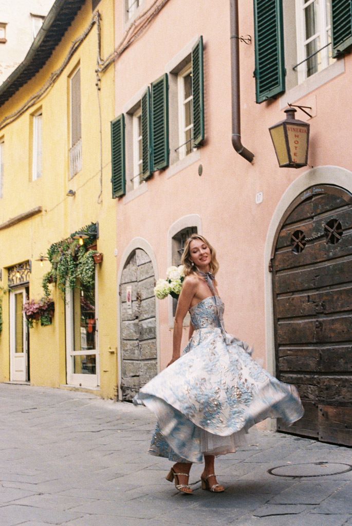 Woman in a blue floral dress twirling on a cobblestone street with colorful buildings in the background during engagement session in Lucca, Italy
