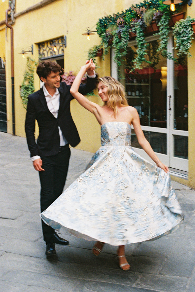 A couple dances joyfully in a charming cobblestone street lined with flowers and colorful buildings during their Lucca, Italy engagement session