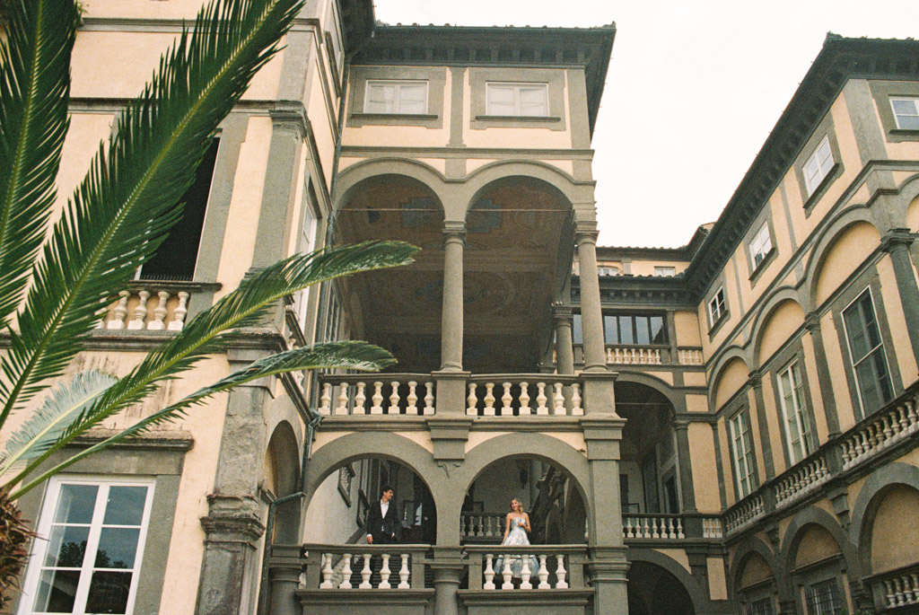 Film photo of couple standing on a grand balcony of an elegant, historic building with arched columns in Lucca, Italy