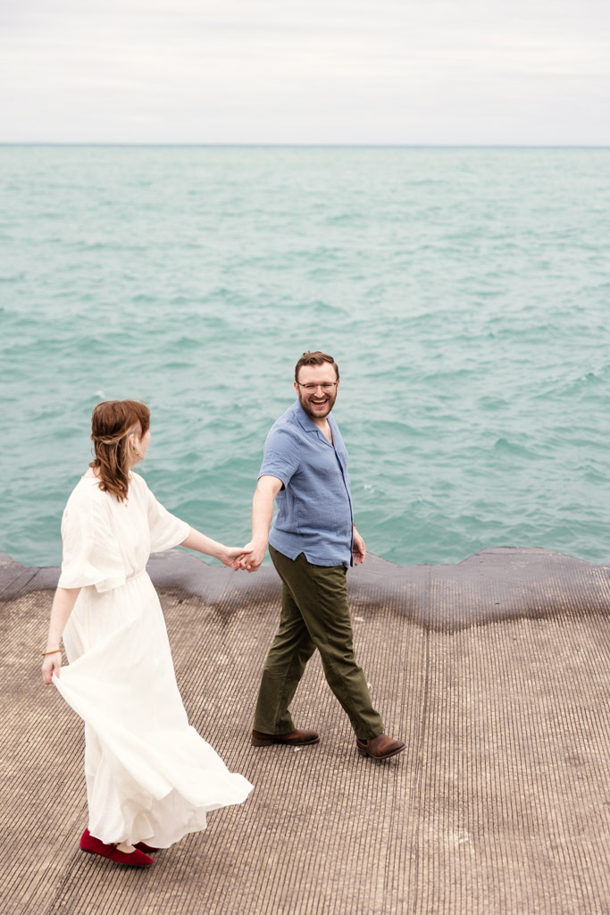 A couple holding hands walks on a pier by the water, smiling and looking at each other, reminiscing about their recent bookstore engagement.