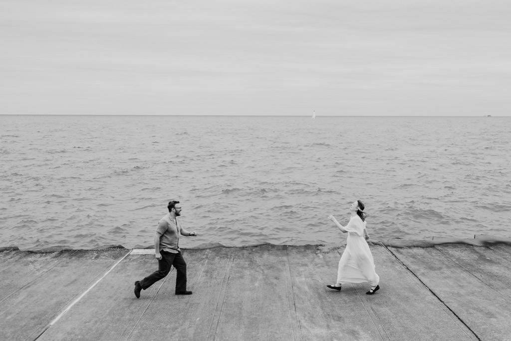 Two people joyfully run toward each other on a concrete path by the sea, sky overcast, their recent bookstore engagement adding an extra spark to their smiles.