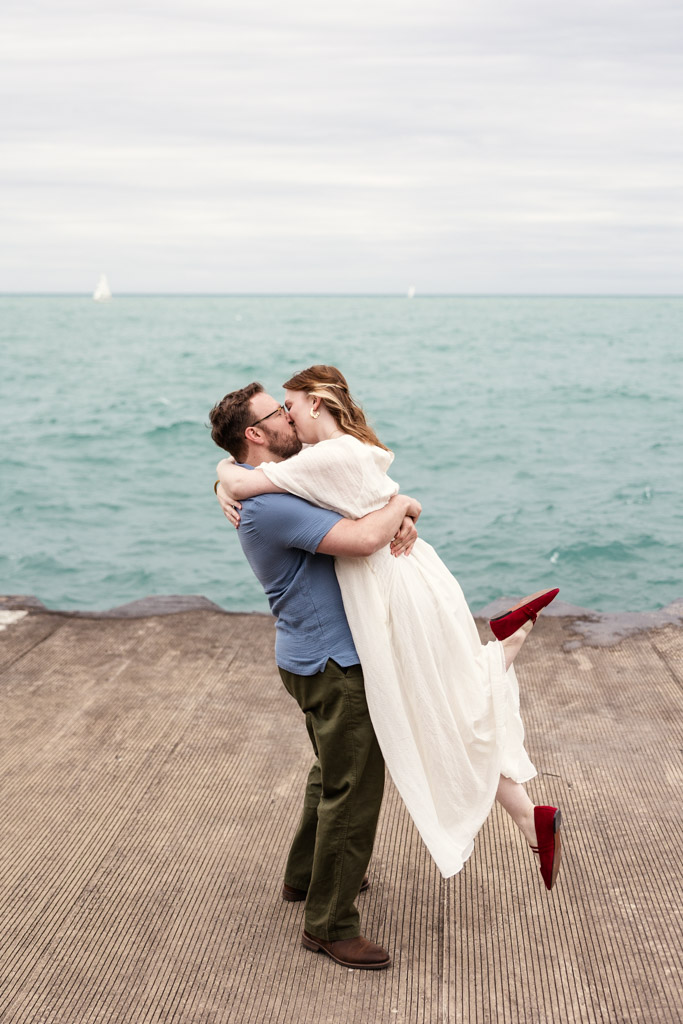 A man lifts and hugs a woman in a white dress by the water, both smiling on a cloudy day, their joy reminiscent of their bookstore engagement.