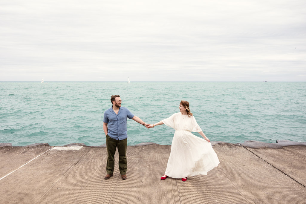 A couple holds hands and faces each other on a pier by the water under a cloudy sky, their smiles reflecting the joy of their recent bookstore engagement.