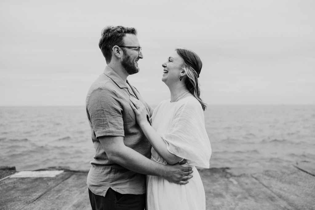 A couple stands by the water, smiling and laughing together on a cloudy day, reminiscing about their recent bookstore engagement.