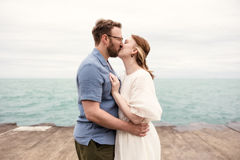 A couple stands by the water, embracing and sharing a kiss on a cloudy day, their love story unfolding like a bookstore engagement—intimate, heartfelt, and timeless.