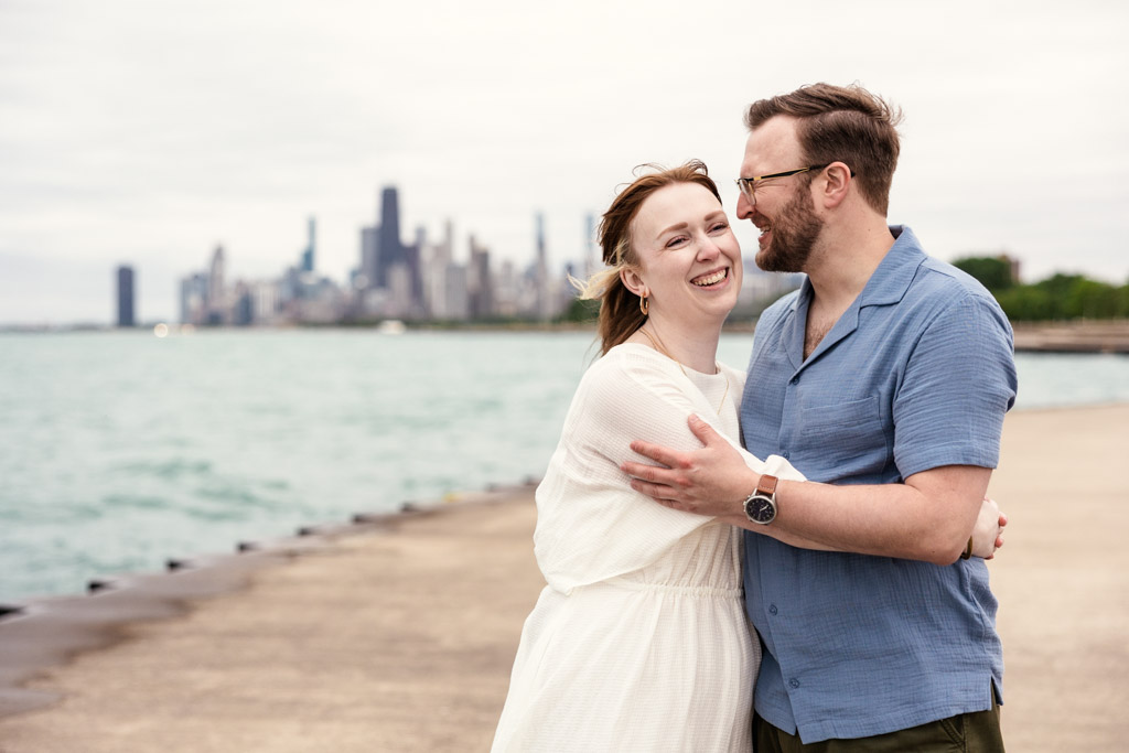 Smiling couple embraces by the waterfront with a city skyline in the background on a cloudy day, sharing a quiet moment reminiscent of their bookstore engagement.