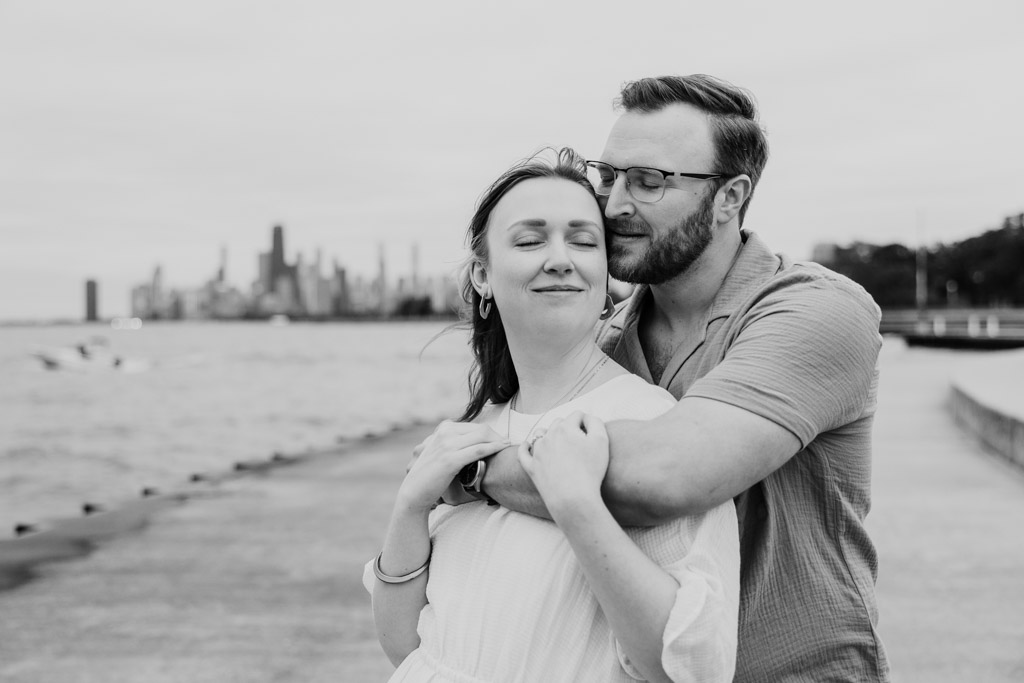 A couple shares a tender embrace on a waterfront path with a city skyline in the background, their love story reminiscent of a perfect bookstore engagement.