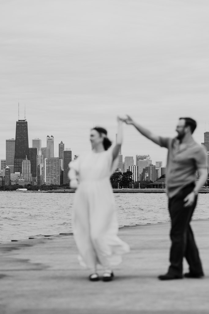 A couple dances near the waterfront with a city skyline in the background, their bookstore engagement adding a charming touch to the scene, though the image is out of focus.