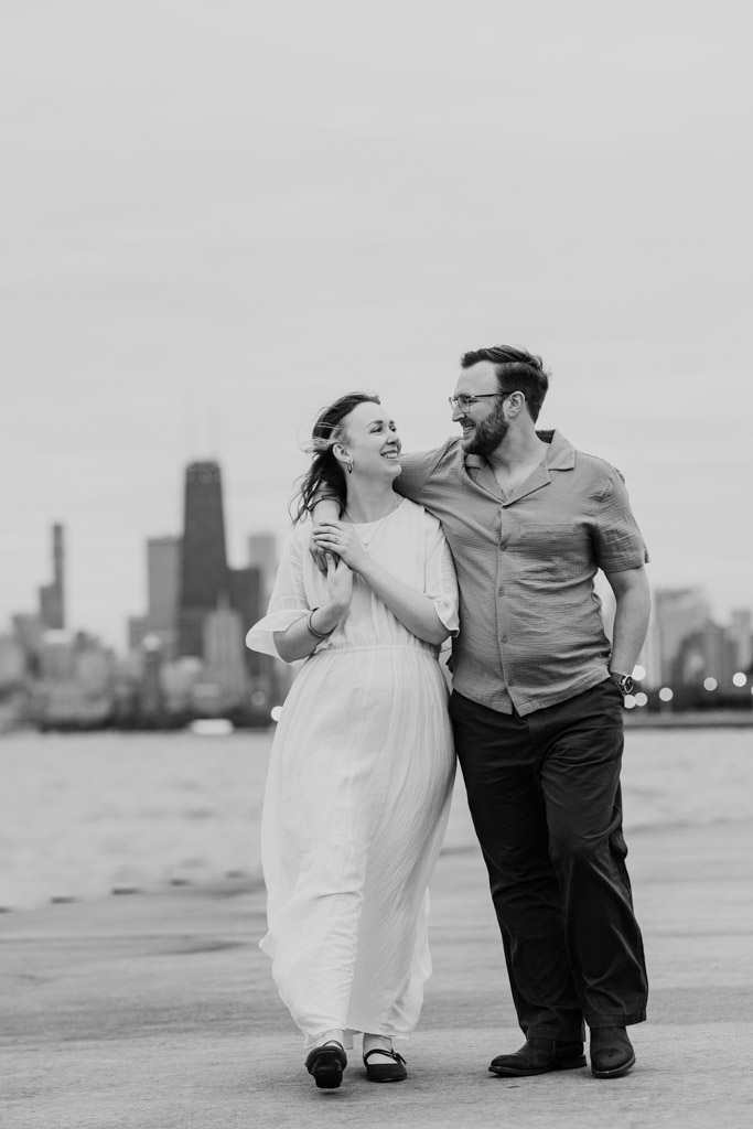 A couple smiles and walks together near water, city skyline in the background, in black and white, capturing the same warmth as their recent bookstore engagement.