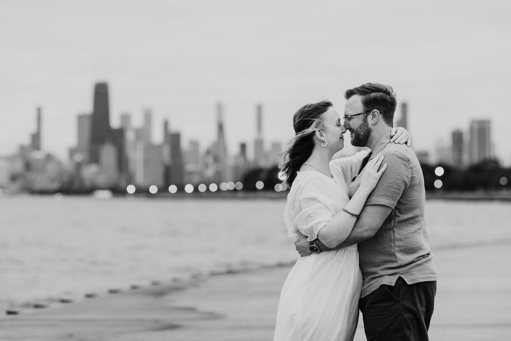 A couple embraces and smiles on a waterfront with a city skyline in the background, radiating the same warmth and joy as they did during their bookstore engagement.