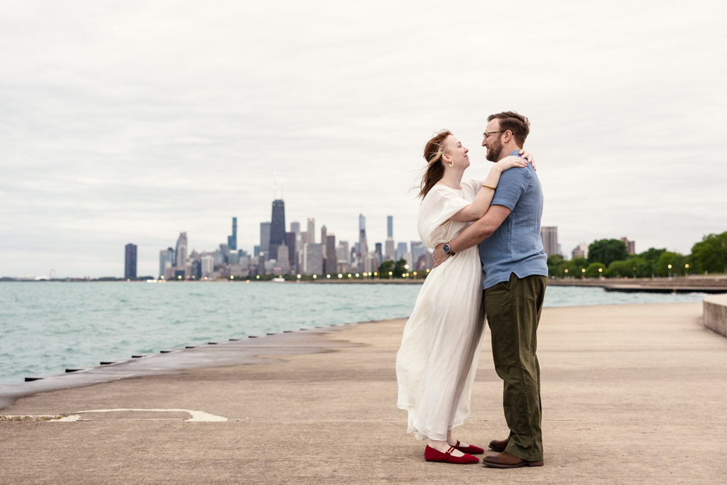 A couple embraces on a waterfront promenade with a city skyline in the background, capturing the warmth of their bookstore engagement amidst an urban romance.