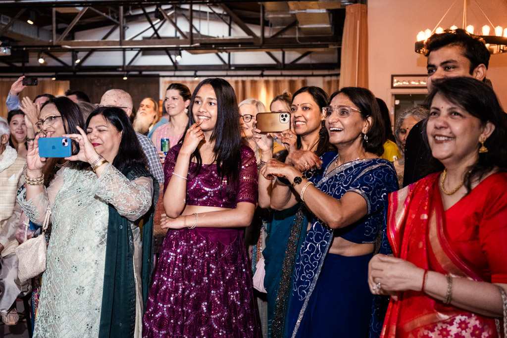 A joyful group of people in colorful outfits smile and take photos at a lively indoor Chicago Sangeet celebration.