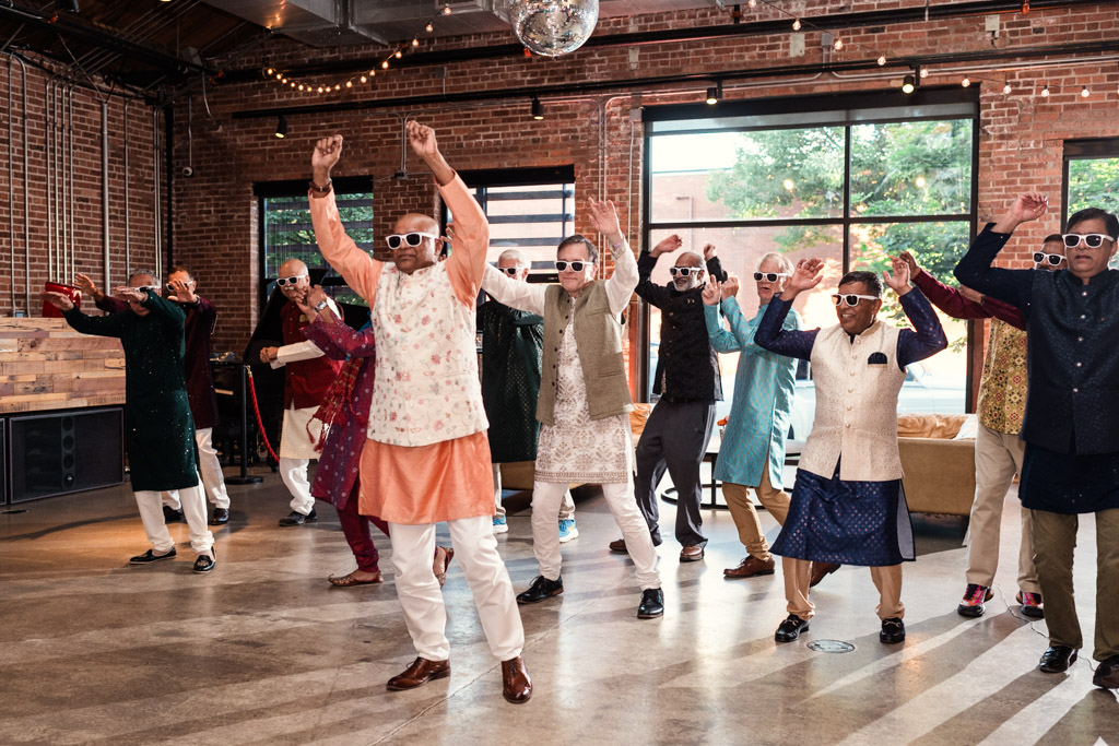 A group of men in colorful traditional outfits dance indoors at a lively Chicago Sangeet, all wearing white sunglasses and smiling.