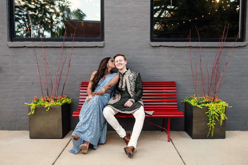Bride kisses groom on the cheek while they sit on a red bench outside Ovation Chicago, dressed in traditional Indian attire