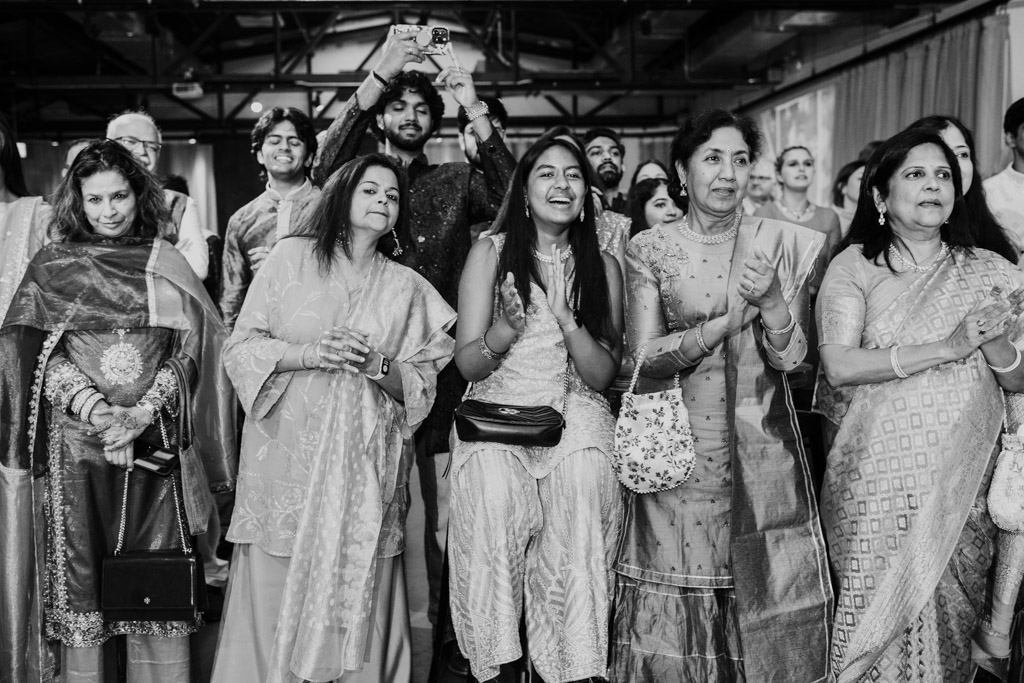 A group of people dressed in traditional attire are smiling and clapping at a lively Chicago Sangeet event.