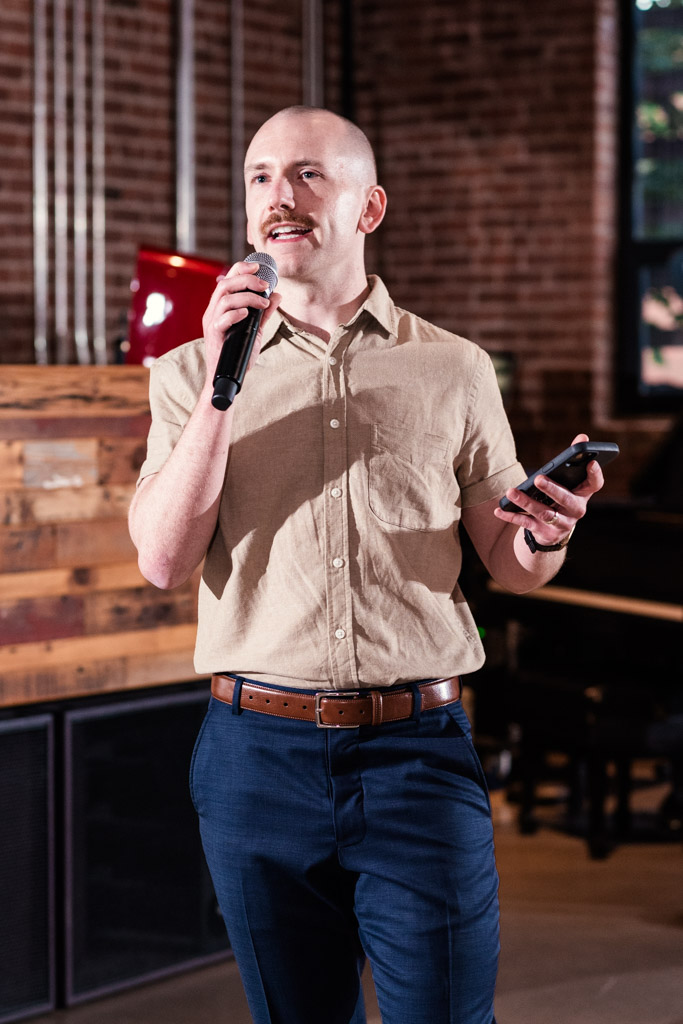 Man in a tan shirt and blue pants speaks into a microphone while holding a phone, indoors with brick walls, at a lively Chicago Sangeet celebration.