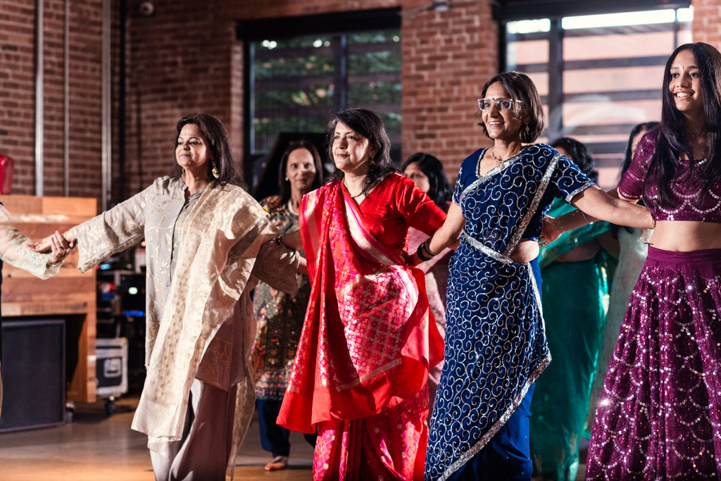 A group of women in colorful traditional clothing dance together indoors, smiling and holding hands as they celebrate a joyful Chicago Sangeet.