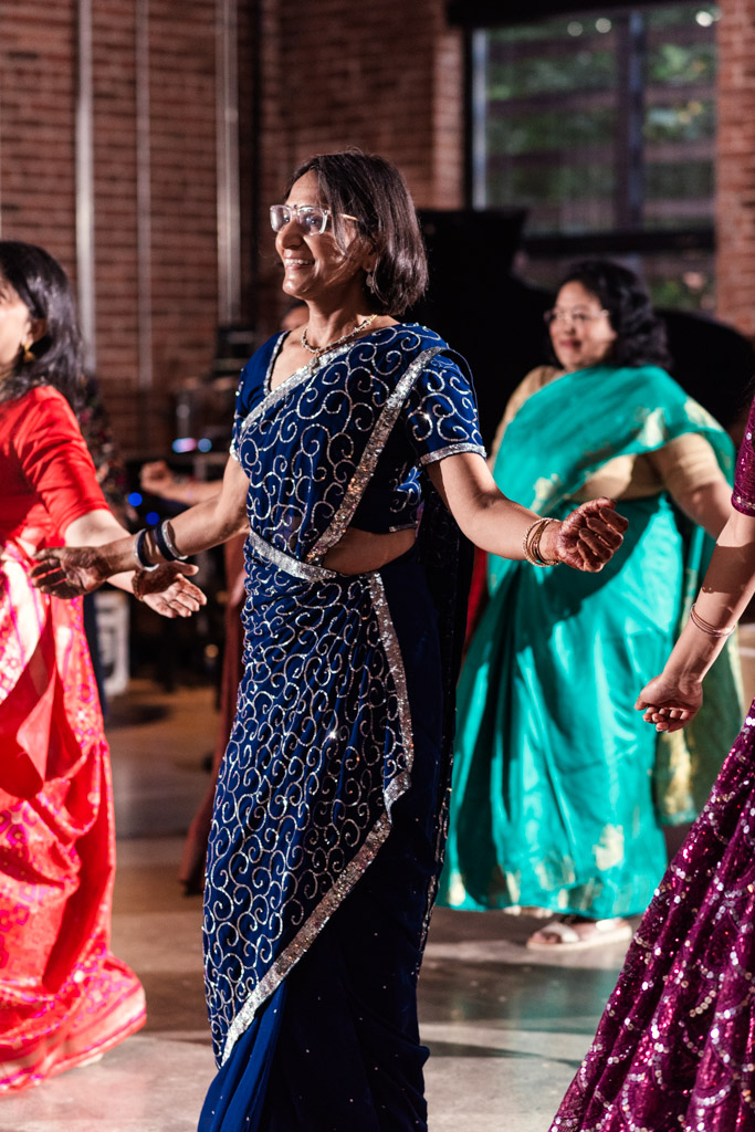 Women in colorful sarees dance together indoors at a lively Chicago Sangeet, smiling and enjoying the event.
