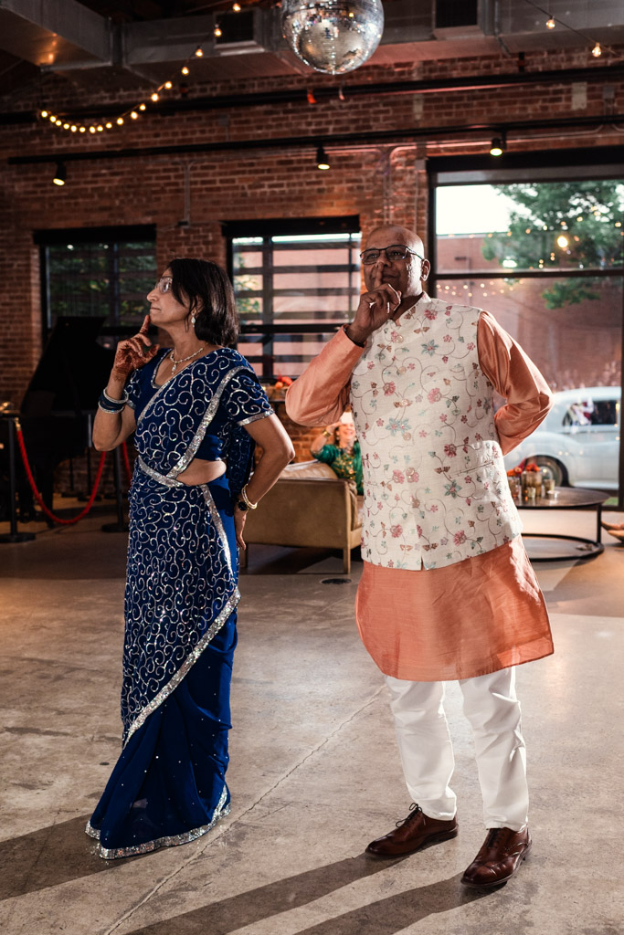 Two adults in formal attire stand indoors, both looking left and touching their faces thoughtfully, as if deep in conversation at a Chicago Sangeet.
