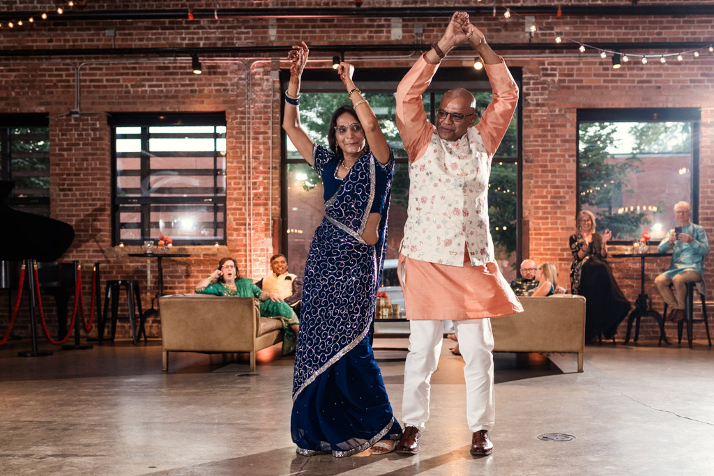A couple in traditional Indian attire dances joyfully in a spacious, brick-walled event hall during their lively Chicago Sangeet.