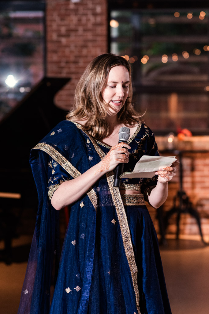 Woman in a blue traditional dress smiles while speaking into a microphone and holding a piece of paper at the Chicago Sangeet.