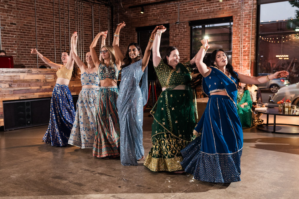 Six women in colorful traditional outfits dance joyfully indoors, arms raised and smiling, capturing the vibrant spirit of a Chicago Sangeet in a festive setting.