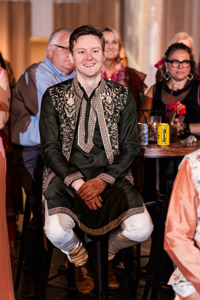 Smiling man in ornate dark green traditional attire sits on a tall stool among a group of people at a lively Chicago Sangeet event.
