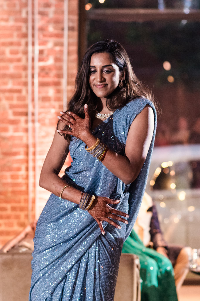 Woman in a sparkly blue sari dancing at a Chicago Sangeet, with henna on her hands, in a warmly lit indoor setting.