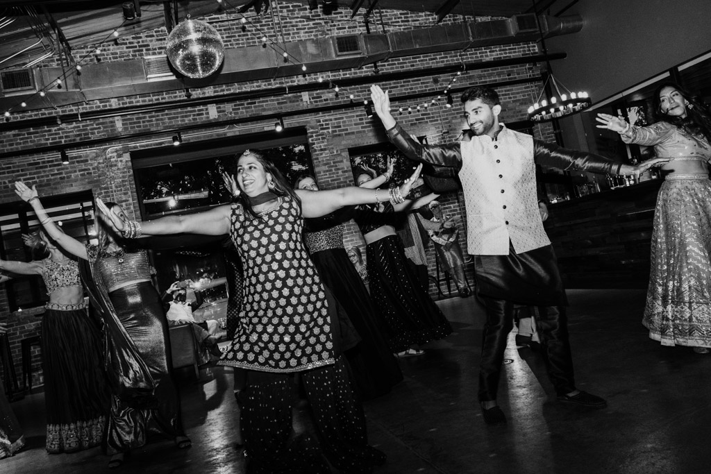 A group of people in traditional outfits dance joyfully at an indoor Chicago Sangeet event under a disco ball.