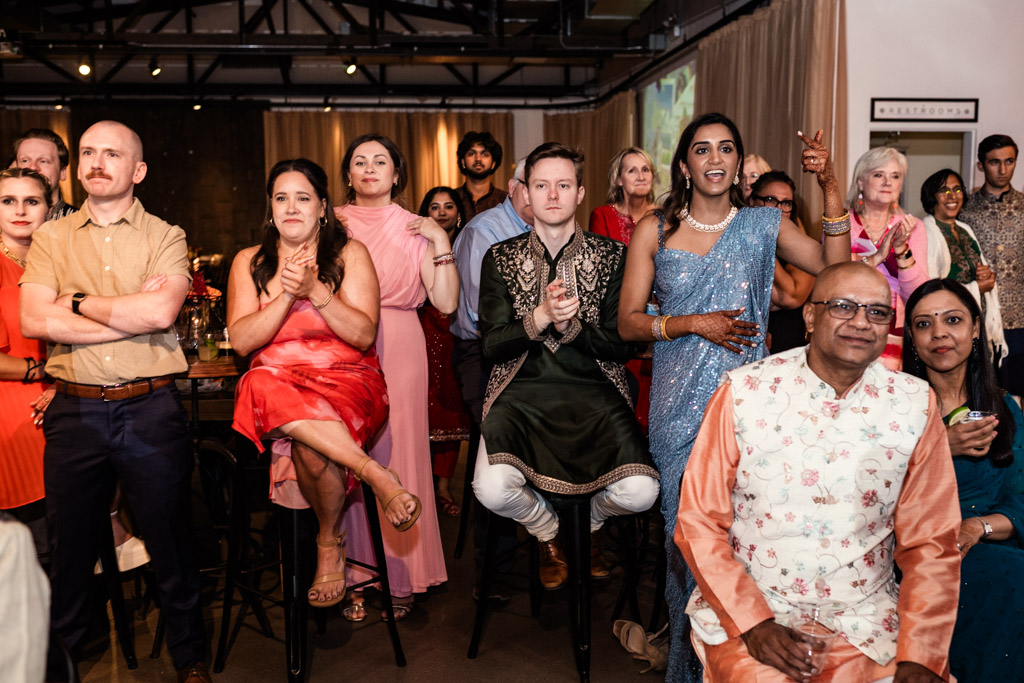 A diverse group of people in colorful attire watch a Chicago Sangeet event indoors, appearing engaged and cheerful.