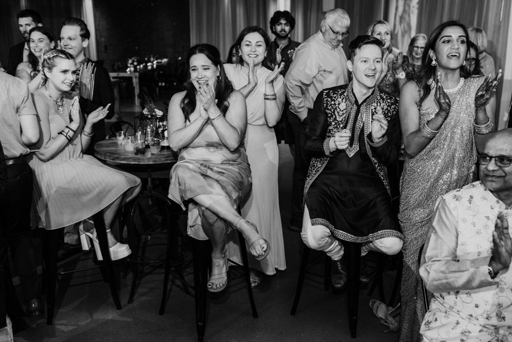 A group of people dressed formally sit and stand, clapping and smiling at a Chicago Sangeet event.