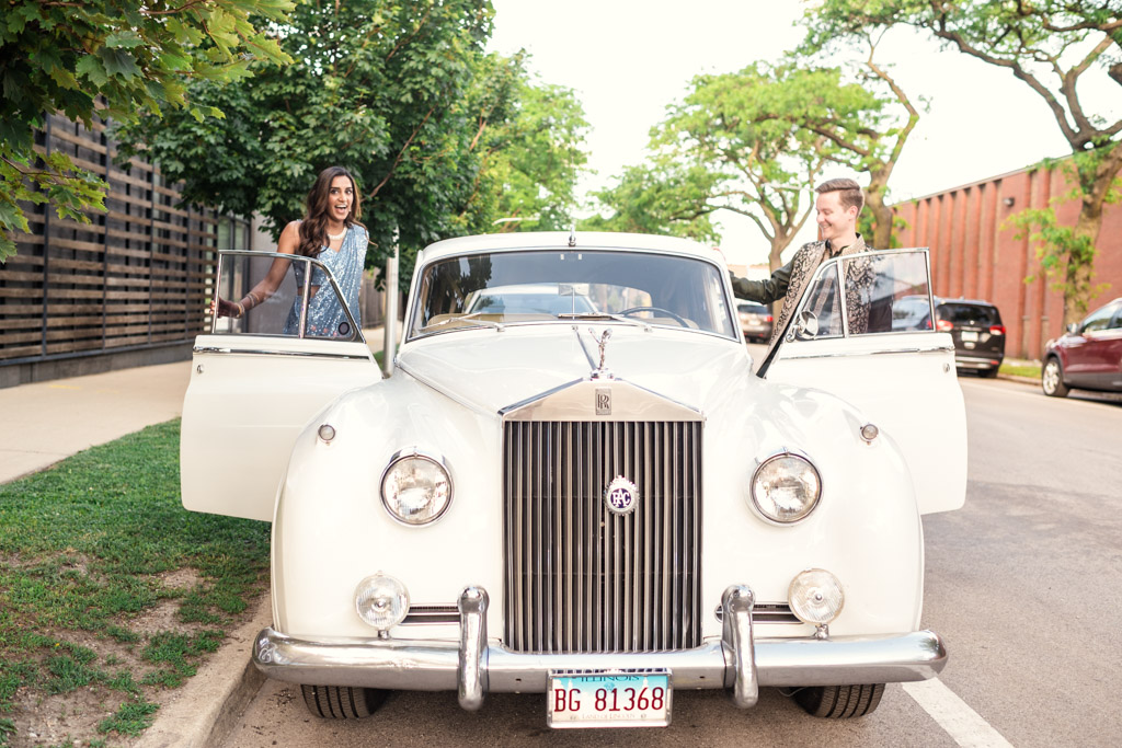 Bride and groom get into a white vintage car parked outside Ovation Chicago on a sunny street with trees and buildings during their Sangeet
