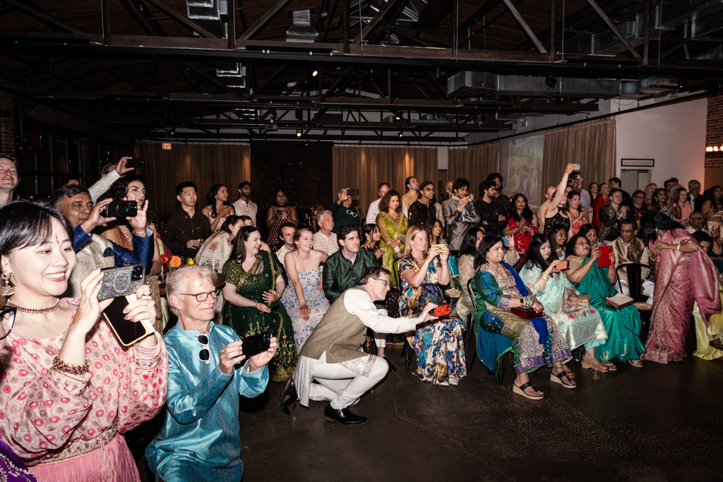 A large group of people in colorful attire watches and takes photos at an indoor event or celebration, capturing the vibrant energy of a Chicago Sangeet.