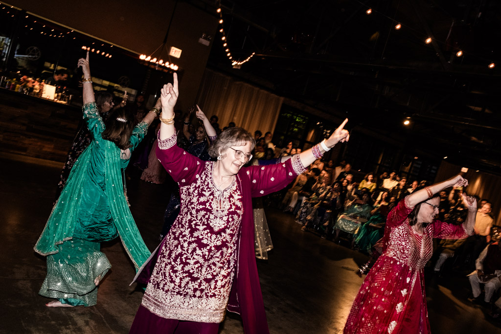 People in colorful traditional clothing joyfully dancing at an indoor Chicago Sangeet event, with string lights twinkling above.