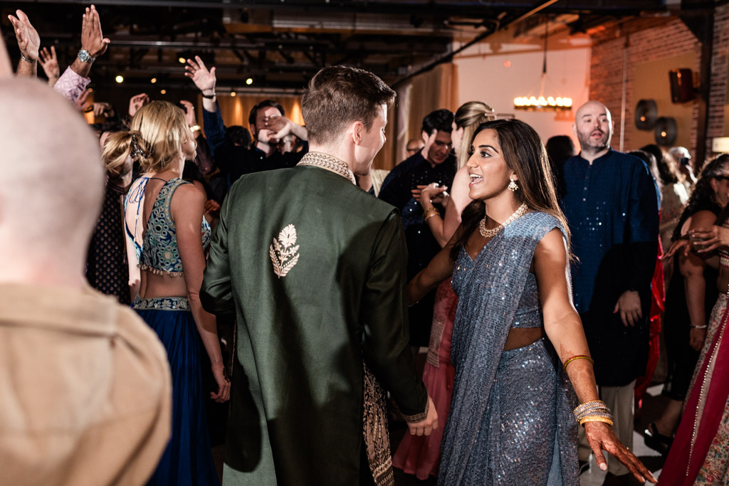 A couple in traditional attire dances joyfully at a lively Chicago Sangeet celebration, surrounded by others joining in the festivities.