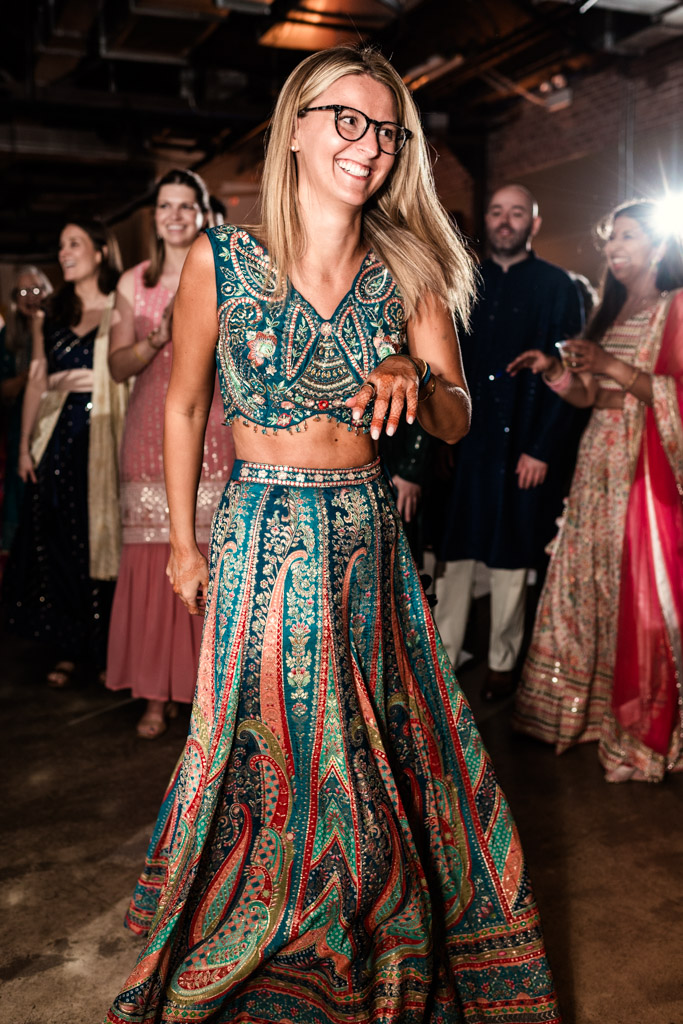 Smiling woman in a colorful lehenga dances at an indoor Chicago Sangeet event, with people in festive attire in the background.