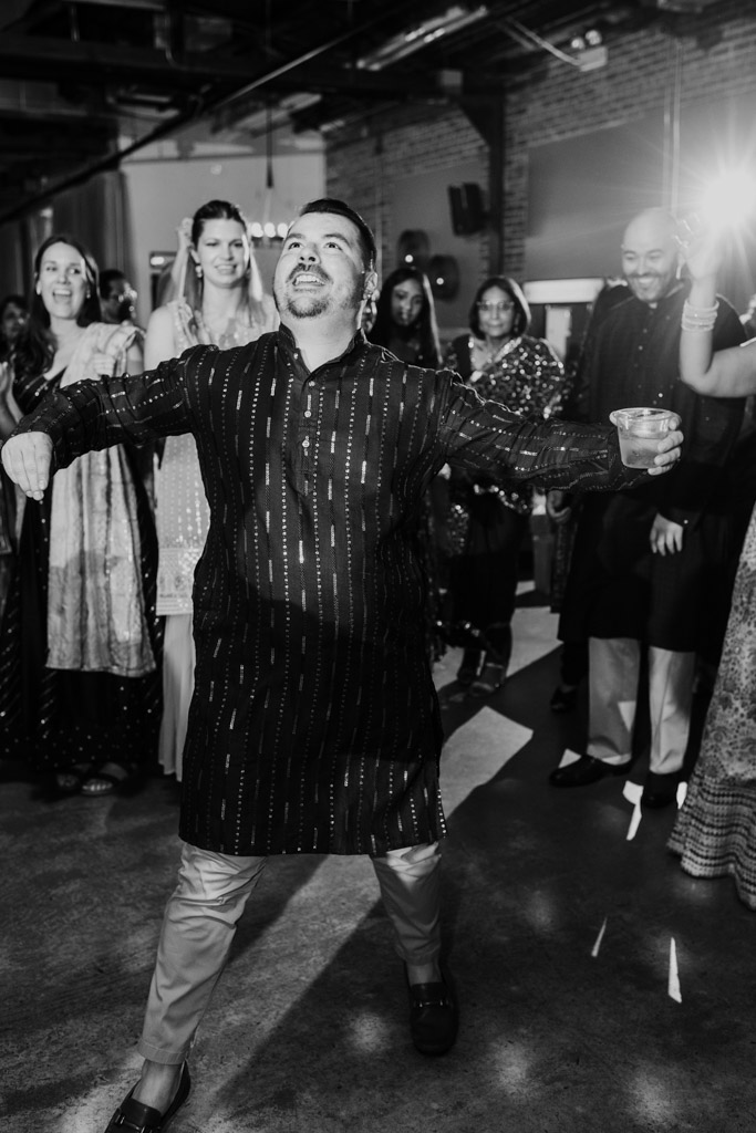 Man dancing joyfully at a Chicago Sangeet, holding a drink, surrounded by smiling people in festive attire.