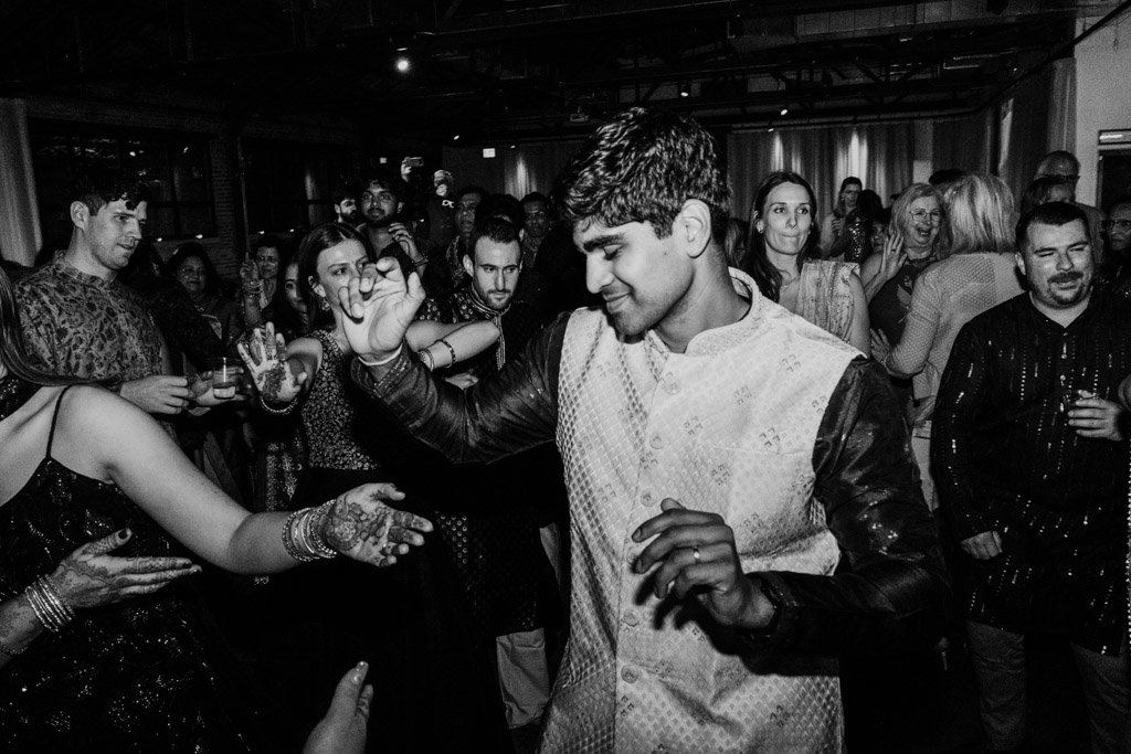 A man in traditional attire dances joyfully at a crowded indoor celebration, embodying the vibrant spirit of a Chicago Sangeet.