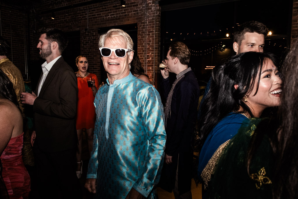 Older man in sunglasses and blue kurta smiles at indoor Chicago Sangeet party with people chatting around him.