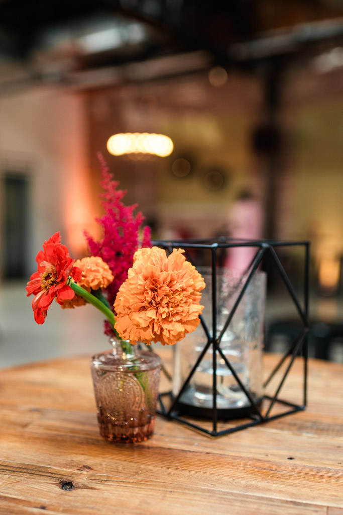 Small glass vase with orange flowers beside a geometric candle holder on a wooden table for Sangeet at Ovation Chicago