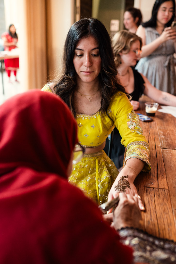 Woman in a yellow dress gets henna applied to her hand at a lively indoor Chicago Sangeet gathering.